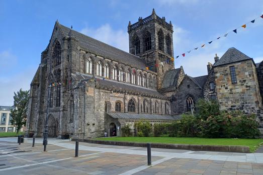 Image of a large grey stone abbey with a large central tower, blue skies, green grass and colourful bunting.