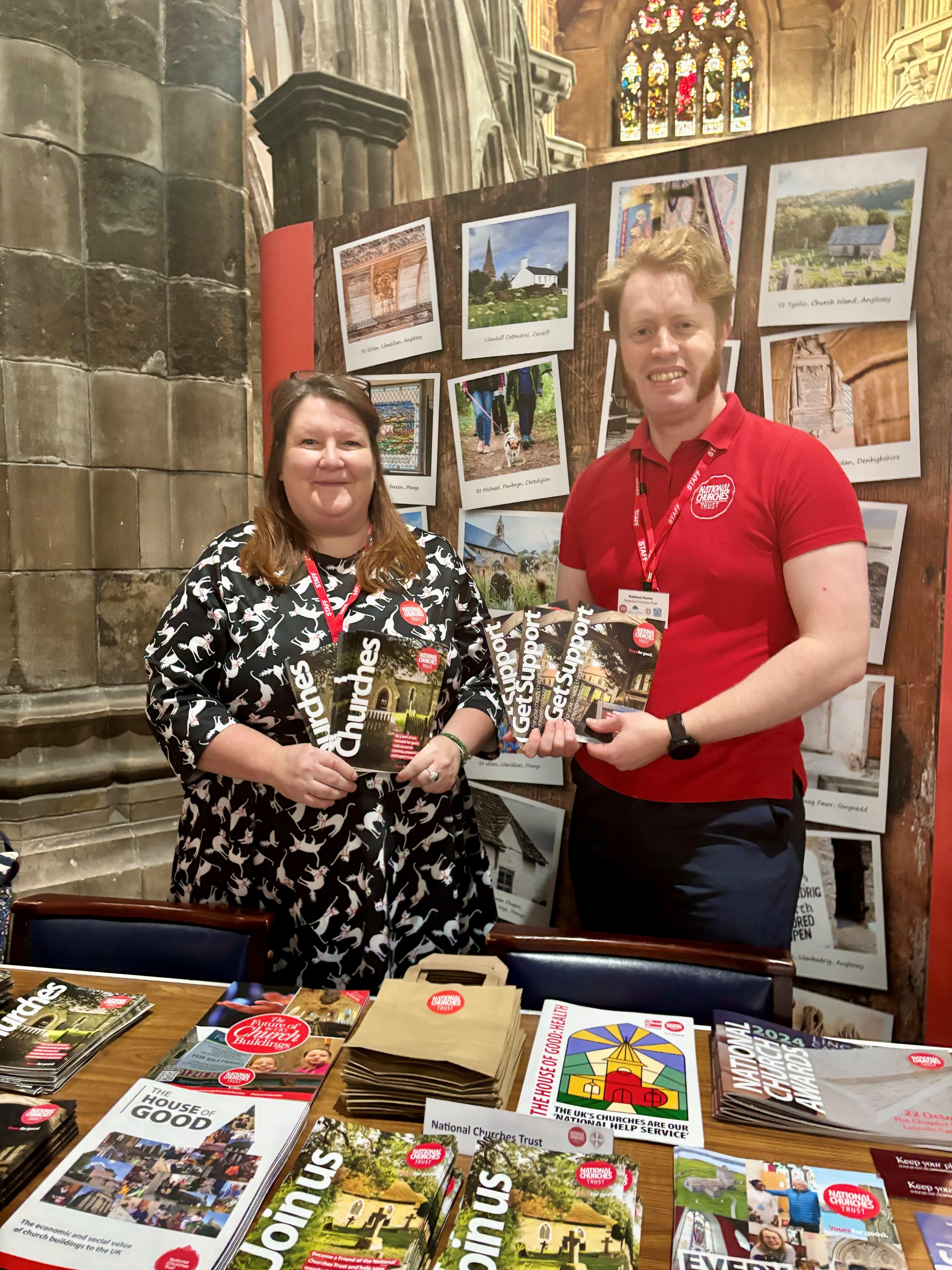 A man and a woman at an event stall, each holding up leaflets