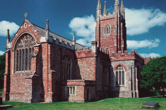 Exterior shot of Totnes St Mary. Red bricked with large arched windows. In the background is a bright blue sky.