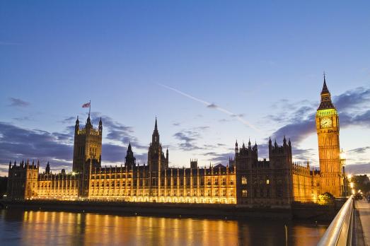 The UK Houses of Parliament lit up with a blue sky behind and part of the river Thames visible 